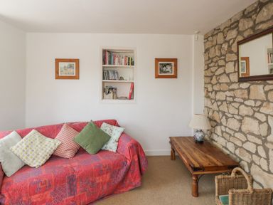 A sitting area with a red covered sofa with cushions a wooden coffee table with a lamp a wicker basket and a stone wall with a mirror at Athelstan Cottage in Chloe Mead near Stroud