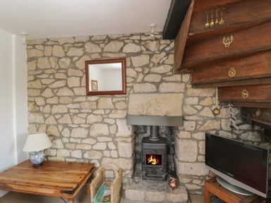 A room with a stone fireplace and wood burning stove a wooden table with a lamp and a television at Athelstan Cottage Chloe Mead near Stroud