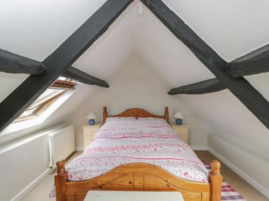 An attic bedroom with a wooden bed a floral bedspread two bedside tables with lamps and exposed dark ceiling beams at Athelstan Cottage in Chloe Mead near Stroud