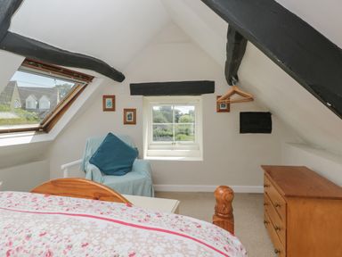 A bedroom with a bed a wooden dresser a chair with a cushion and two windows at Athelstan Cottage in Chloe Mead near Stroud