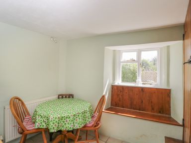 A small dining area with a round table covered by a green floral tablecloth two wooden chairs and a wooden window seat at Athelstan Cottage in Chloe Mead near Stroud