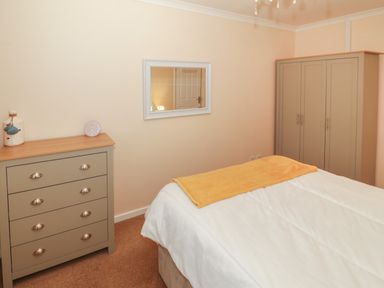 A bedroom with a bed covered with white bedding and an orange blanket a grey dresser with a clock and vase a grey wardrobe and a wall mirror at Glanaig Morfa Bychan