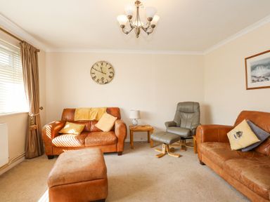 A living room with two brown leather sofas a matching ottoman a green recliner chair a wooden side table with a lamp a wall clock curtains and a framed picture at Glanaig Morfa Bychan