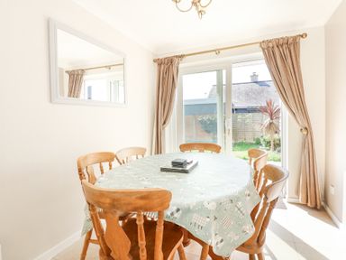 A dining room with a round table covered by a patterned tablecloth and six wooden chairs near sliding glass doors with curtains at Glanaig Morfa Bychan