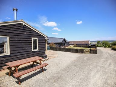 An outside area with a wooden picnic table beside a black wooden building and a gravel driveway at Norton Cottage in Craven Arms