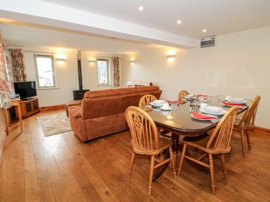 A dining area with a wooden table set with plates and red napkins next to a living room with brown sofas a rug a small television and two windows at Norton Cottage in Craven Arms