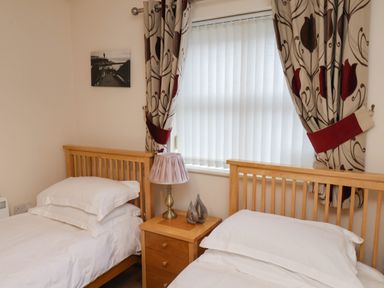 A bedroom with two single wooden beds a wooden nightstand with a lamp and decor between them and a window with vertical blinds and curtains at Cadliam in Whitby