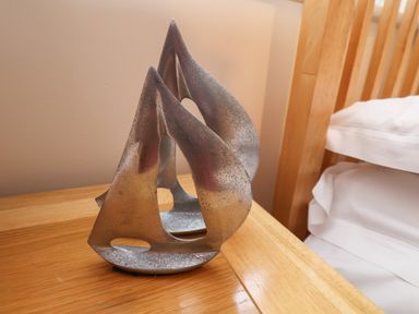 A small metal sailboat sculpture on a wooden bedside table next to a bed with white bedding at Cadliam in Whitby