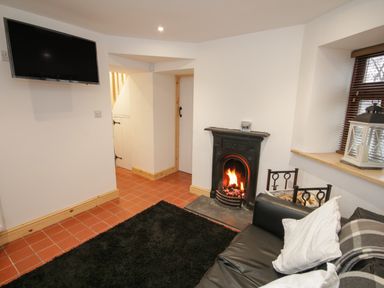 A living room with a black sofa white pillows a fireplace wall mounted television and a window with blinds at Old Tollgate in Gwalchmai near Llangefni