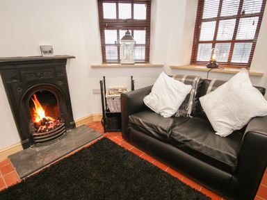 A room with a black leather sofa with white pillows a fireplace with a fire and windows with wooden blinds at Old Tollgate in Gwalchmai near Llangefni