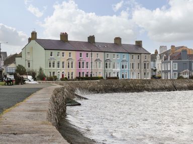 A row of colorful terraced houses along a waterfront with a stone seawall and benches at Old Tollgate in Gwalchmai near Llangefni