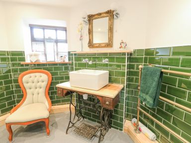 A bathroom with green tiled walls a vintage sewing machine table sink an upholstered chair and a towel on a rail at Old Tollgate in Gwalchmai near Llangefni