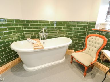 A bathroom with a white freestanding bathtub green tiled walls and a cushioned wooden chair at Old Tollgate in Gwalchmai near Llangefni