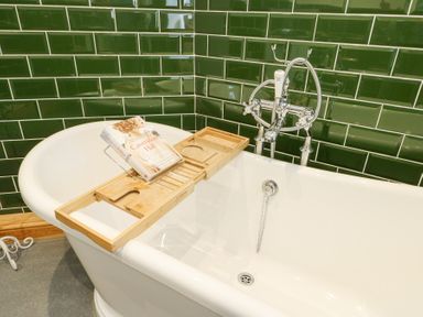 A white bathtub with a wooden bath tray holding a book and green tiled walls in the bathroom at Old Tollgate in Gwalchmai near Llangefni