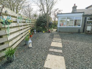A gravel garden with a stone pathway leading to a house with a white conservatory and plants along a wooden fence at Old Tollgate in Gwalchmai near Llangefni