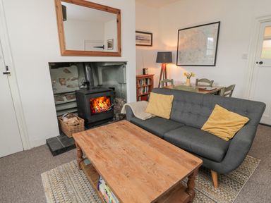 A living room with a wood stove, a grey sofa with yellow cushions, a wooden coffee table, a dining table with chairs, a floor lamp, and framed pictures on the walls at Lavender Cottage in Glanton