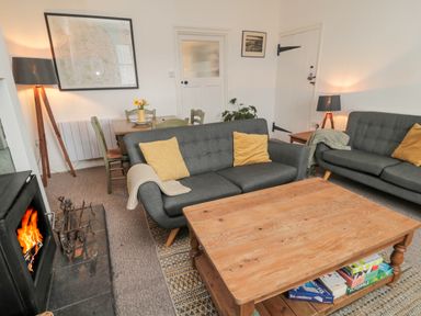A living room with gray sofas yellow cushions a wooden coffee table board games under the table a fireplace a dining table with green chairs and a floor lamp at Lavender Cottage in Glanton