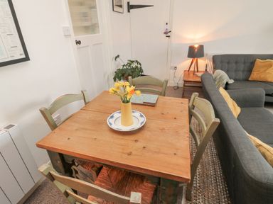 A dining area with a wooden table and chairs and flowers in a vase next to a living room with a gray sofa and a table lamp at Lavender Cottage in Glanton