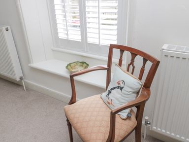 A wooden chair with a puffin cushion near a window with white shutters and a radiator at Lavender Cottage in Glanton