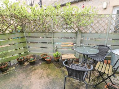 A small outdoor patio with a round glass table two wicker chairs one wooden chair several potted plants and a green wooden fence at Lavender Cottage in Glanton