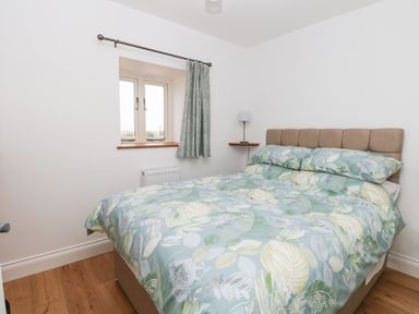A bedroom with a double bed with patterned bedding and a window with a curtain at Waveney View Cottage in Burgh Castle