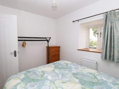 A bedroom with a bed covered by a blanket with leaf patterns a wooden chest of drawers a metal clothing rack with hangers a window with blue-green curtains and a door at Waveney View Cottage in Burgh Castle