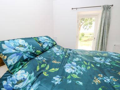A bedroom with a bed covered in blue floral bedding near a window with curtains at Waveney View Cottage in Burgh Castle