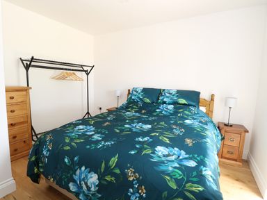 A bedroom with a double bed with floral bedding wooden side tables lamps a chest of drawers and a clothing rack with hangers at Waveney View Cottage in Burgh Castle