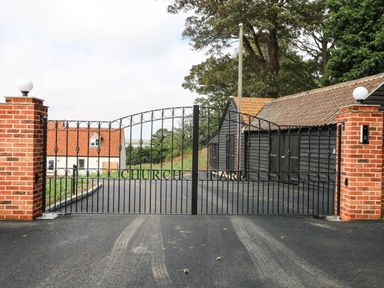 A black metal gate with brick pillars at the entrance of a driveway leading to a white house and black outbuildings at Church Farm in Burgh Castle