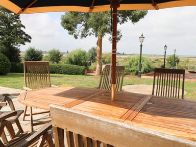 A wooden outdoor dining table with chairs under an umbrella in a garden with trees and lamp posts at Waveney View Cottage in Burgh Castle