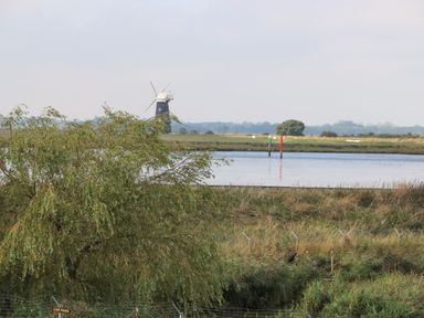 A river with a windmill and navigation markers in the background at Waveney View Cottage in Burgh Castle