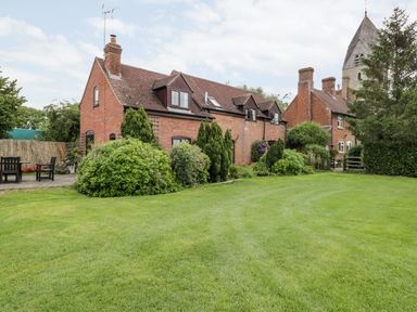 A brick house with dormer windows surrounded by bushes and a lawn with outdoor chairs at Lawn Farm Cottage in Churcham near Gloucester