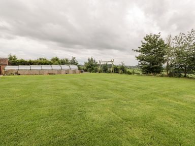 A large grassy lawn with a covered swimming pool and a wooden swing set at Lawn Farm Cottage in Churcham near Gloucester