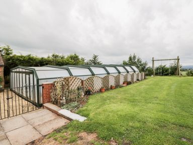 A garden area with a large greenhouse, lattice fencing, potted plants, and a swing set at Lawn Farm Cottage in Churcham near Gloucester