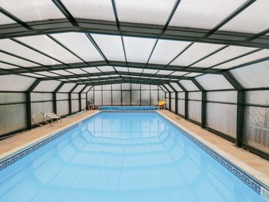 An indoor swimming pool enclosed by a glass and metal frame structure with chairs and pool equipment at Lawn Farm Cottage in Churcham near Gloucester