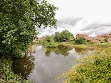 A pond surrounded by trees bushes and grass with brick houses in the background at Lawn Farm Cottage in Churcham near Gloucester