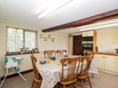 A dining area with a table covered in a polka dot tablecloth and wooden chairs near a window with lace curtains and a high chair at Lawn Farm Cottage in Churcham near Gloucester