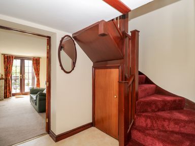 A hallway with a wooden staircase with red carpet and a small door underneath next to a round mirror and an adjacent room with a sofa and glass doors at Lawn Farm Cottage in Churcham near Gloucester