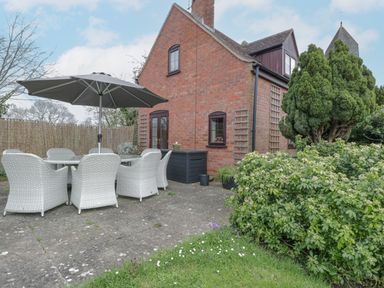 An outdoor patio area with a table and chairs under a large umbrella beside a brick house with greenery and bushes at Lawn Farm Cottage in Churcham near Gloucester