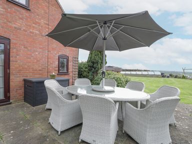 An outdoor patio with a glass table and six white wicker chairs under a large gray umbrella at Lawn Farm Cottage in Churcham near Gloucester
