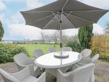 An outdoor patio set with a glass top table umbrellas and chairs on a stone surface with grass and trees in the background at Lawn Farm Cottage in Churcham near Gloucester