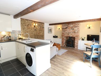 An open kitchen and living area with a washing machine a brick fireplace and a small dining table at Captain's Cottage in Sheringham