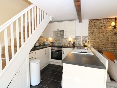 A kitchen with white cabinets a black countertop patterned tile backsplash a staircase and a stone wall at Captain's Cottage in Sheringham