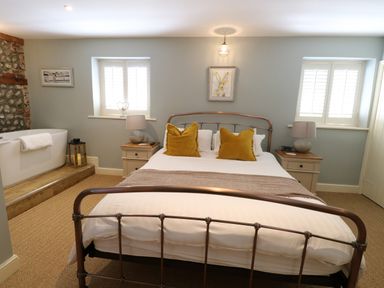 A bedroom with a bed with yellow pillows two bedside tables and a bathtub near a stone wall at Captain's Cottage in Sheringham
