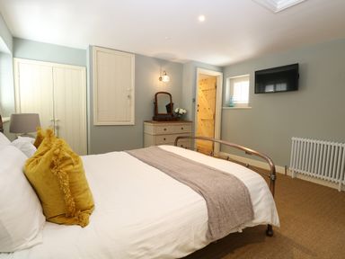 A bedroom with a bed and yellow cushions a dresser with a mirror a wall-mounted TV and a wooden door at Captains Cottage in Sheringham