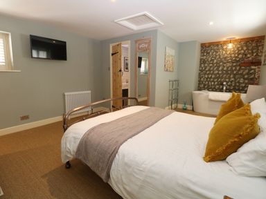 A bedroom with a bed a wall-mounted tv a full-length mirror and a bathtub against a stone wall at Captain's Cottage in Sheringham