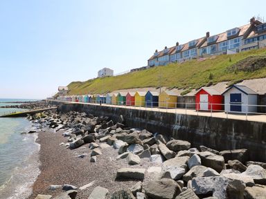 A rocky beach with colorful beach huts along a promenade and houses on a hill at Captains Cottage in Sheringham
