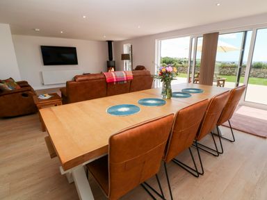 A dining area with a wooden table and brown chairs next to a living room with brown sofas and a wall-mounted TV at Atlantic View in Germoe