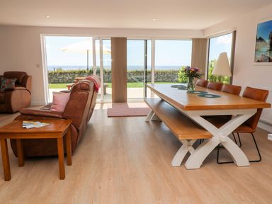 A dining area with a wooden table and leather chairs next to a sitting area with brown leather sofas overlooking a garden and sea at Atlantic View in Germoe