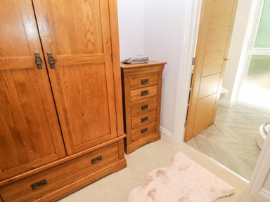 A wooden wardrobe a wooden chest of drawers an open door leading to a bathroom with tiled floor and a pink rug on carpeted floor at Atlantic View in Germoe